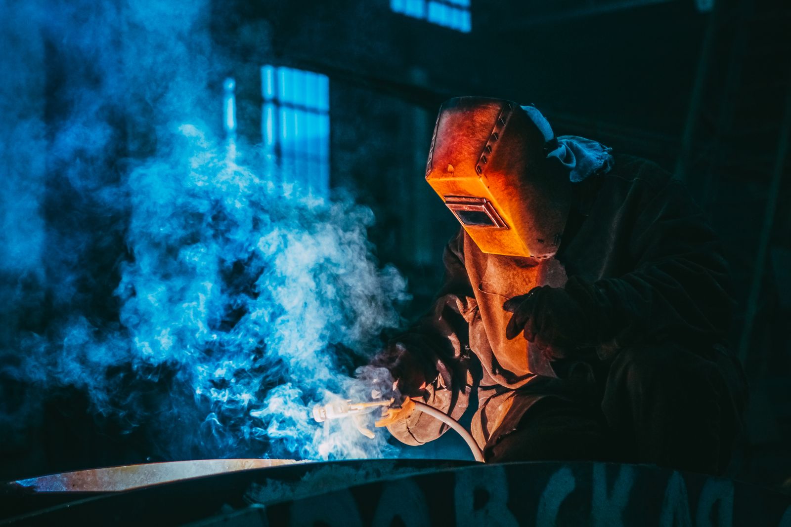 A welder in protective gear at work, sparks and smoke lit against a dark workshop