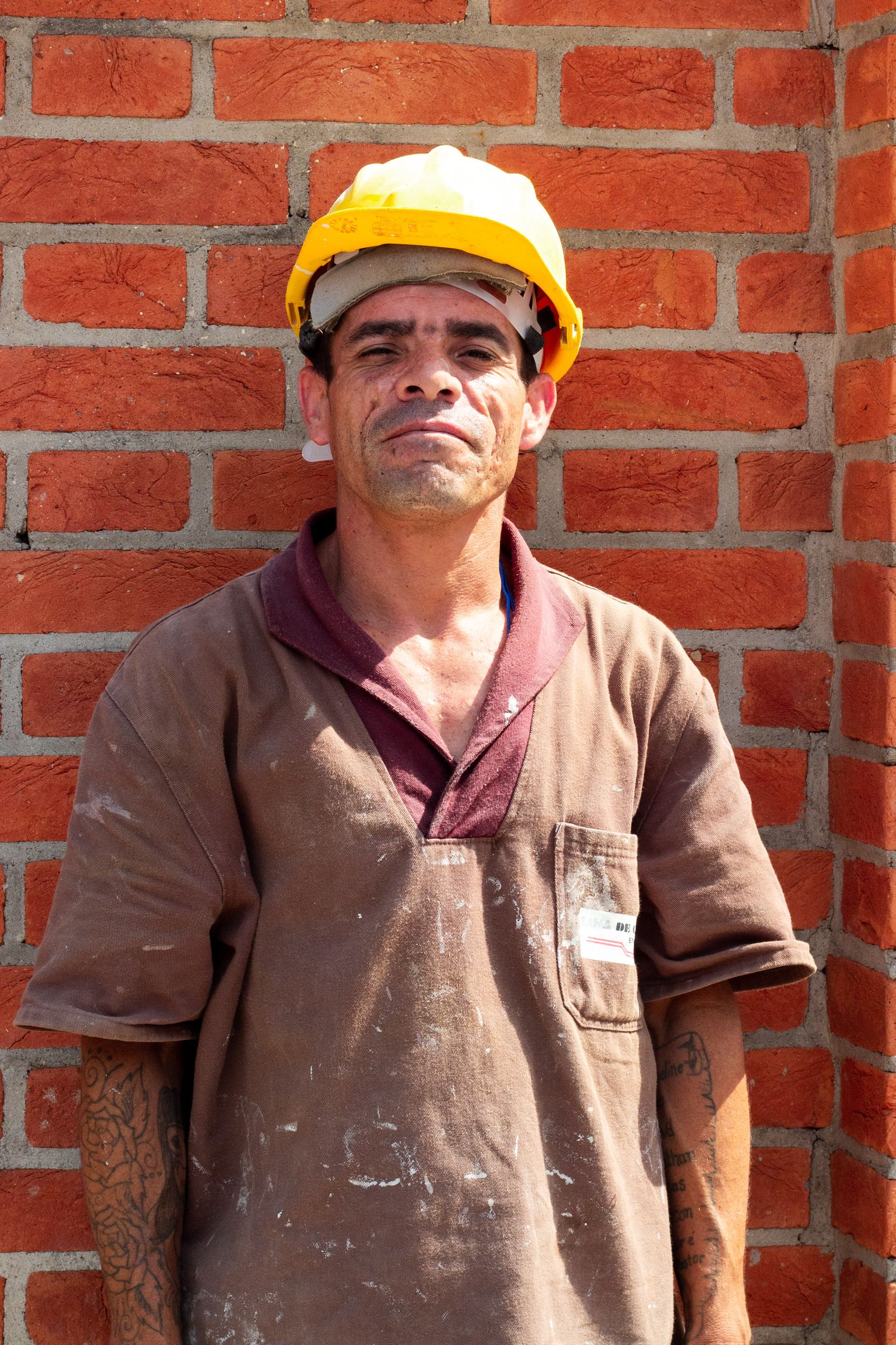 A tradesperson in worn overalls standing against a red brick wall